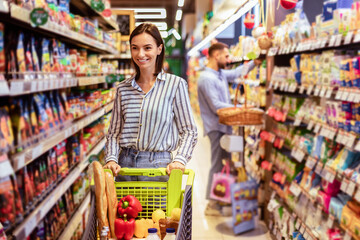 Cheerful Young Lady Doing Shopping Buying Groceries In Supermarket Standing And Pushing Trolley Cart Full Of Healhty Food, Smiling Woman Choosing Products And Looking At Shelf Indoors