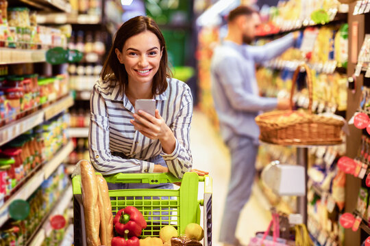 Portrait of smiling young casual woman leaning on shopping trolley cart indoors, using her mobile phone, posing and looking at camera. Satisfied female customer buying products in local grocery store