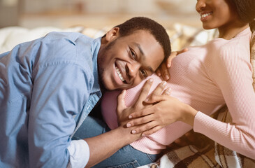 Happy black man hugging his pregnant girlfriend's big belly and listening to unborn baby's...