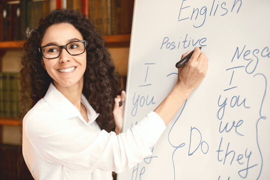 Lecture. Closeup portrait of cheerful smiling female tutor wearing glasses teaching English language, writing grammar rules on the board with marker, looking back at her students at classroom