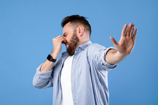 Bad smell concept. Young bearded guy closing his nose and showing STOP gesture, expressing disgust on blue studio background. Millennial man repulsed by toxic odor or terrible perfume