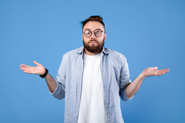 Fototapeta premium Who knows. Confused bearded man in glasses shrugging shoulders on blue studio background. Unsure millennial guy expressing uncertainty, feeling clueless, making hard choice