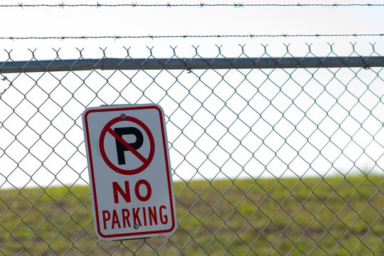 A white metal sign with a red round circle and the letter P in black paint. The signage is on a metal chain-link fence. The notice says no parking in red letters. The restricted area has green grass. 