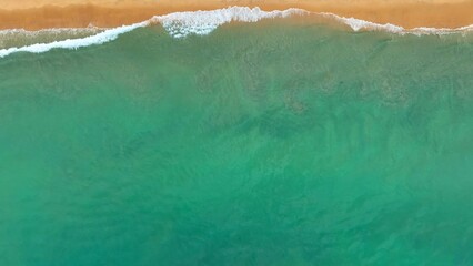 The aerial view captures the mighty ocean as waves crash against the shore, releasing immense power as the water surges over the golden sands in a stunning display of nature’s force.
