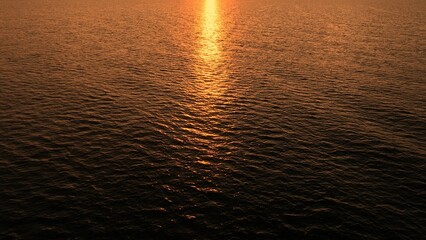 Aerial perspective reveals the shimmering, rippling surface of the sea, illuminated by the setting sun, with the light streaking across the water like liquid gold.
