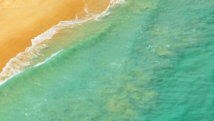 From above, the tropical sea's gentle waves flow toward the golden beach, their foamy crests merging with the calm water, offering a peaceful and picturesque view of nature's beauty.
