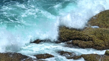 A dramatic close-up shot of ocean waves crashing against rocky shorelines, creating a powerful display of nature’s force with frothy white surf and clear blue water. Phuket, Thailand.
