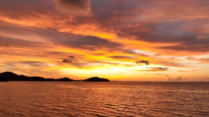 A serene beach sunset, where the sky is ablaze with colors of orange and pink, as the sun sets behind distant hills, casting a warm glow across the peaceful ocean and inviting relaxation. Thailand.
