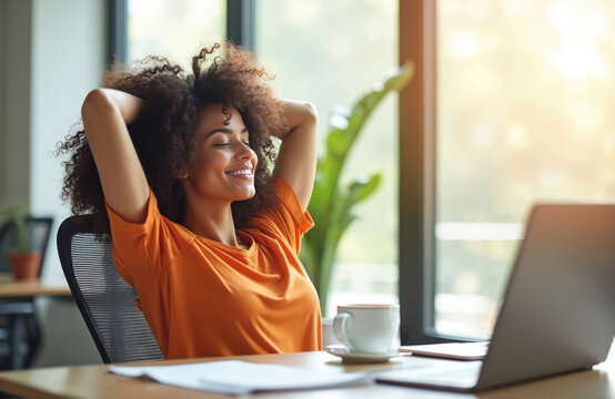 Happy curly-haired woman taking break in modern office, relaxing at desk with cup of coffee, laptop. Smiling female with hands behind head enjoying calm moment. Leisure, well-being, workplace.