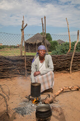 village rural ,portrait of african old woman, cooking cauldron over open fire, in front of a house with thatched roof , countryside living,