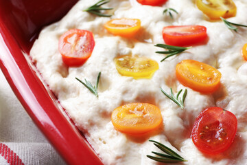 Raw focaccia with tomatoes and rosemary in baking dish on table, closeup