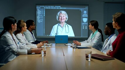 Diverse group of doctors attending an online conference with a senior physician displayed on a large screen, discussing medical cases and treatment strategies in a modern hospital setting - Powered by Adobe