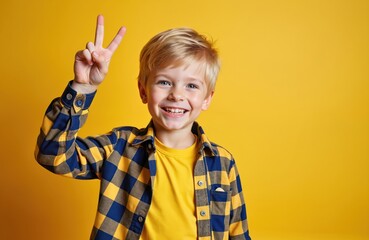 Happy little boy shows victory gesture on yellow background. Smiling kid wearing shirt. Cheerful young male model shows peace fingers symbol. Kid facial expression. Positive emotions, childhood.