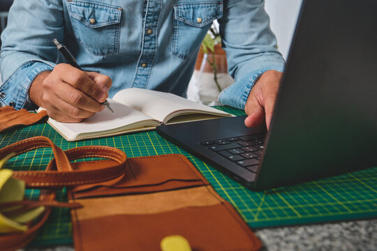 Leather craftsman writing notes and working on laptop, managing his small business from home - Powered by Adobe