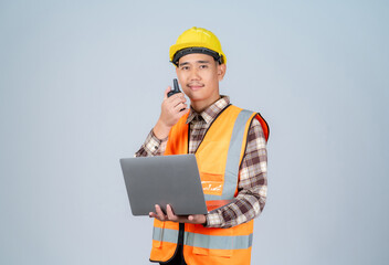 Young Asian engineer or worker wearing yellow hard hat and reflective vest using radio and holding laptop working in studio on grey background scene