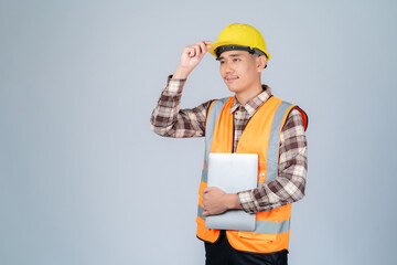 Young Asian engineer or worker wearing yellow hard hat and reflective vest working with laptop in studio on grey background.