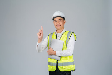 Smiling young Asian engineer or worker pointing upwards and using laptop working in studio on grey background.