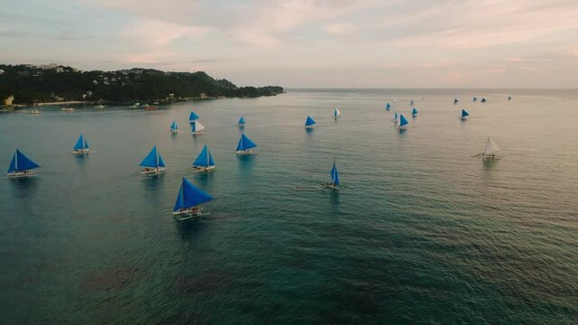Traditional sailboats with blue sails floating on a wide tropical sea under a pastel colored sky. Boracay Island, Philippines.