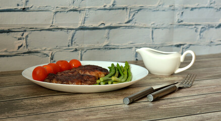 Two pieces of fried beef meat on a plate with cherry tomatoes and asparagus, sauce and cutlery on a light wooden table.