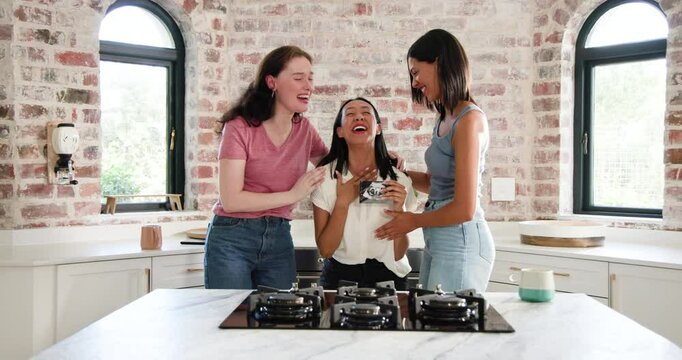 Diverse female friends holding camera and passing card around marble island in kitchen sharing joy