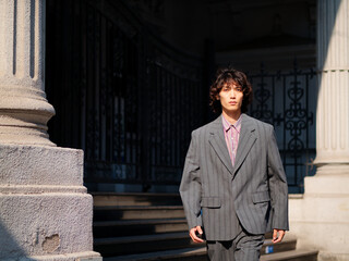 Portrait of handsome Chinese young man wearing gray suit walking in the street, young guy with black curly hair with urban background. Male fashion, cool Asian young man lifestyle.