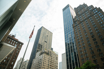 An American flag rises in front of towering buildings in Midtown Manhattan. Glass, brick, and stone architecture represents the city's diverse style.