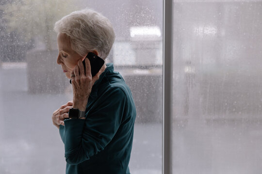 Senior Woman Conversing on Phone Near Window on Rainy Day