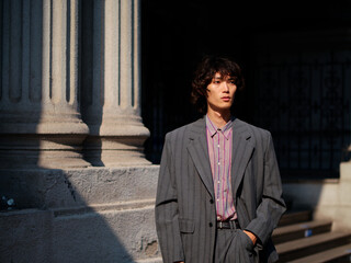 Portrait of handsome Chinese young man wearing gray suit posing in the street, young guy with black curly hair with urban background. Male fashion, cool Asian young man lifestyle.