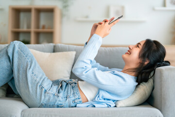 Happy cheerful positive young Asian woman on the phone at home, smiling girl using app and browsing on the internet on smartphone lying on sofa