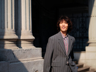Portrait of handsome Chinese young man wearing gray suit posing in the street, young guy with black curly hair with urban background. Male fashion, cool Asian young man lifestyle.