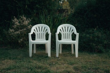 Two white plastic chairs sit on a grassy lawn, facing each other, against a backdrop of lush green foliage.  The scene evokes a quiet, contemplative atmosphere