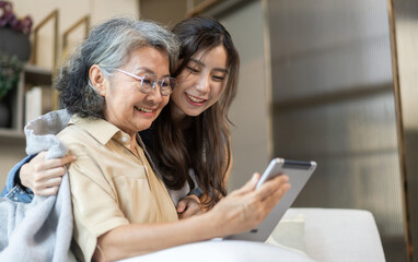 Asian senior mother and daughter smiling and talking using tablet browsing social media, shopping online, watching movies or series, enjoying entertainment digital connection and bonding at home