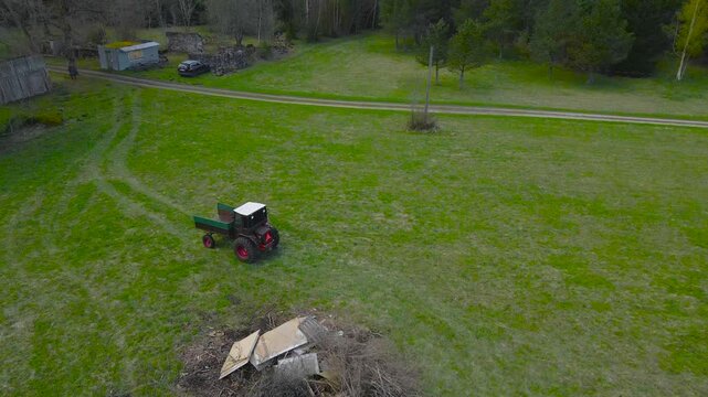 Aerial drone footage flying over and following a retro small vintage green tractor working in a grassy green field where a large pile of garde waste is stored for burning during a cloudy day.
