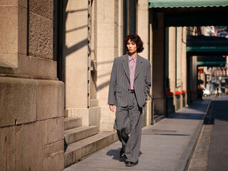 Portrait of handsome Chinese young man wearing gray suit walking in the street, young guy with black curly hair with urban background. Male fashion, cool Asian young man lifestyle.