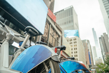 A detailed view of a public bike wheel parked on a New York street. In the background, skyscrapers and LED billboards highlight the city's busy urban style.
