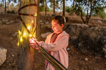 A joyful child wraps colorful string lights around a tree in a serene forest camp, enhancing the natural beauty as evening descends. The atmosphere is warm and inviting for outdoor fun.