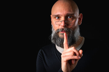 Bearded man making silence gesture with finger on lips. Mysterious secret concept. Portrait on black background with dramatic lighting and serious expression