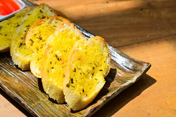 Five slices of garlic bread with fresh parsley on a long plate. Tomato and chilly sauce. Wooden table. Dark, black, shadows, sun light, sunshine. Extreme close up view.