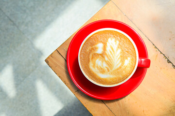 Top view of hot cafe latte in the red cup. Wooden table, grey or gray floor, corner. Copy space. Coffee. Latte art. Close up. High angle, above. Dark, shadows.