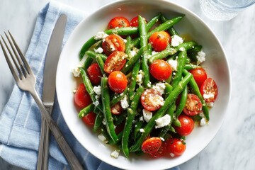 A vibrant green bean and cherry tomato salad, dressed with feta cheese and herbs, is presented in a shallow white bowl.  Cutlery rests on a blue napkin beside it