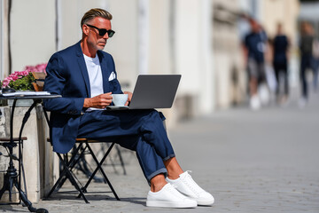 Man in lightweight navy suit with white t-shirt, sitting outside cafe with laptop and espresso, urban summer office look, 