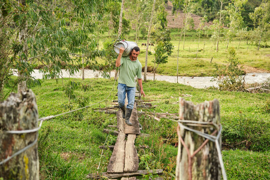 Farmer carrying milk can across wooden bridge in rural landscape