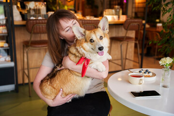 girl dog owner and dog corgi pembroke sitting in dog friendly cafe. on the table is a cup of tea and dessert. girl holds dog in her arms and hugs it