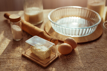 A close-up shows baking ingredients on a textured surface, including butter under a glass dish, two eggs, a rolling pin, and glass jars with flour and sugar.