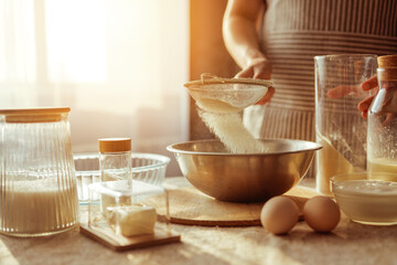 A person wearing an apron sifts flour through a sieve into a metal bowl, surrounded by other baking ingredients on a table near a window.