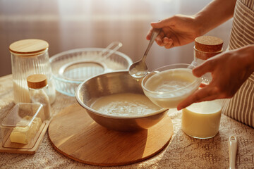 A person in a striped apron adds a creamy mixture from a small glass bowl into a larger metal bowl...