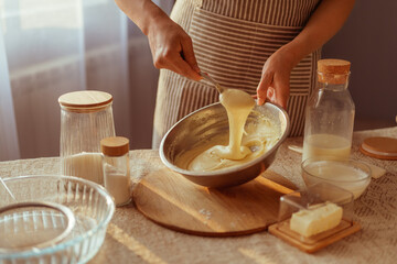 A person wearing a striped apron stirs thick batter in a metal bowl with a spoon, surrounded by...
