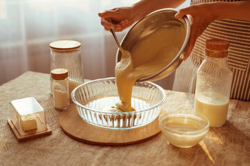 A cook in a striped apron pours thick batter from a metal bowl into a glass pie dish, with other baking ingredients on the table.