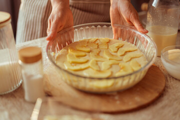 A cook in a striped apron holds a glass pie dish filled with apple slices arranged over batter, ready to be baked.