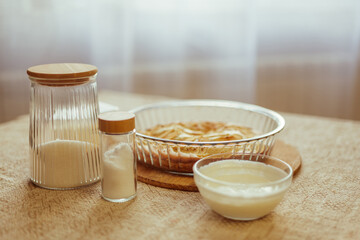 A baked apple pie in a glass dish is on a table with jars of sugar and flour, and a small bowl of creamy liquid.